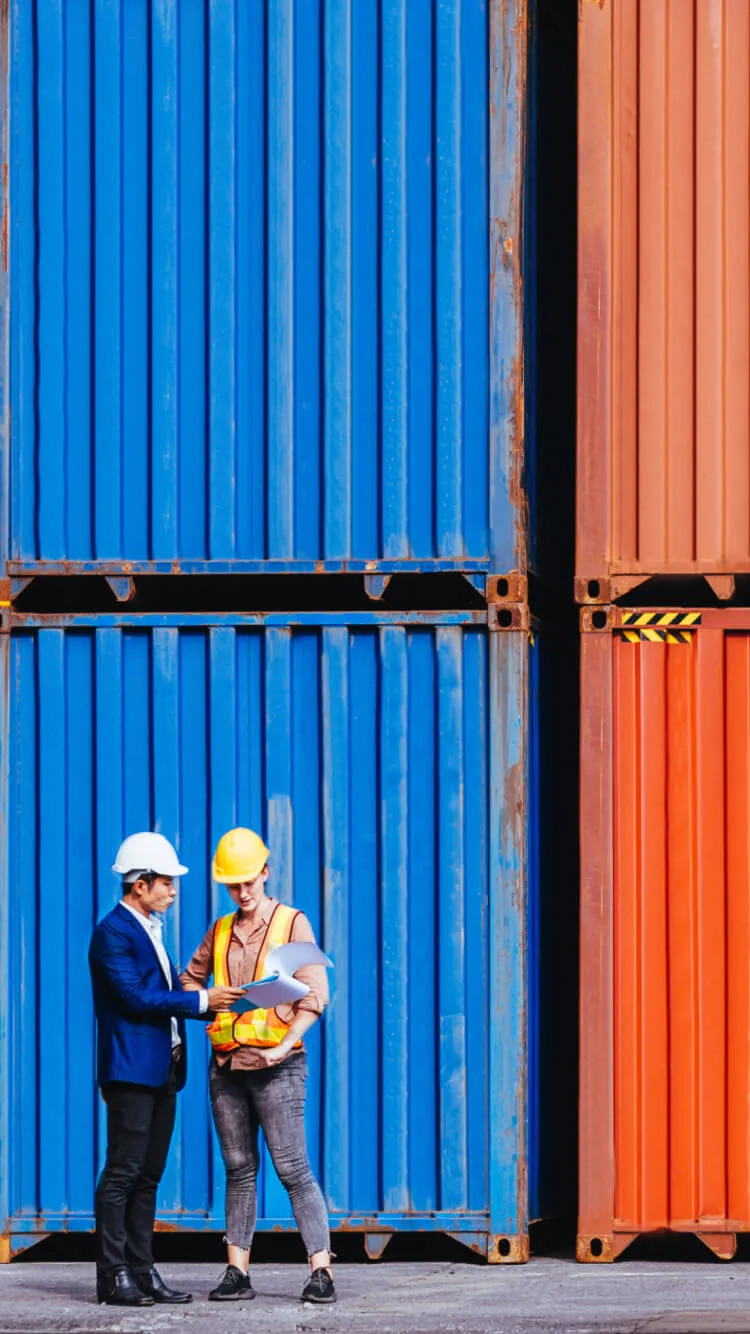 Two workers in hard hats review documents in front of stacked shipping containers.