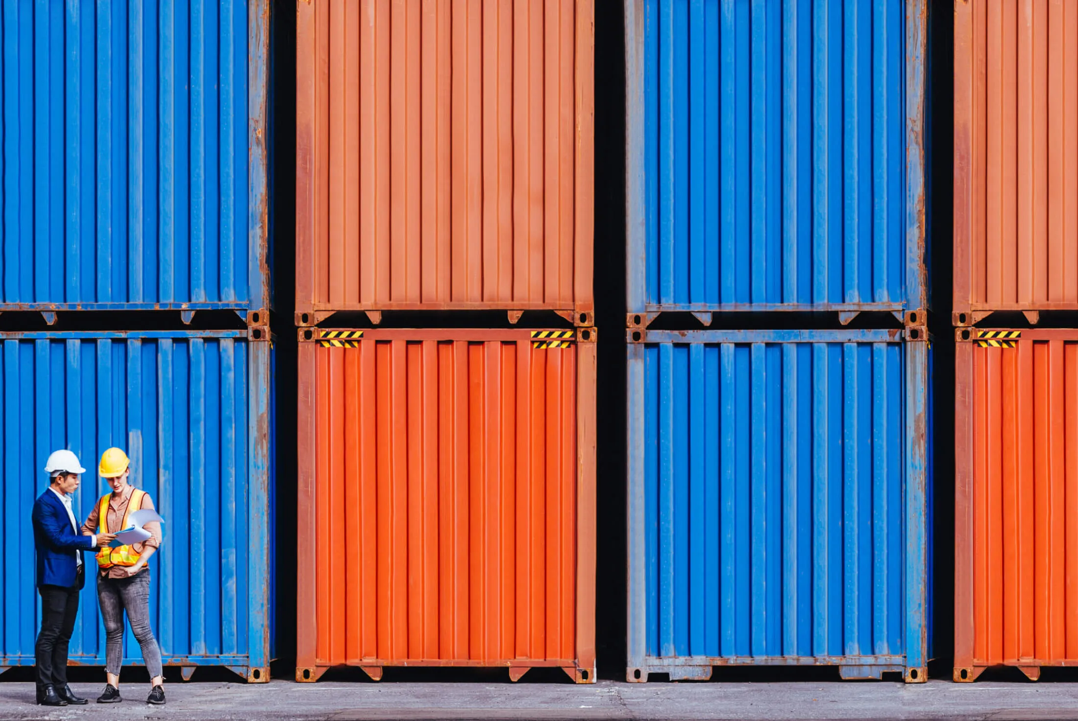 Two workers in hard hats review documents in front of stacked shipping containers.