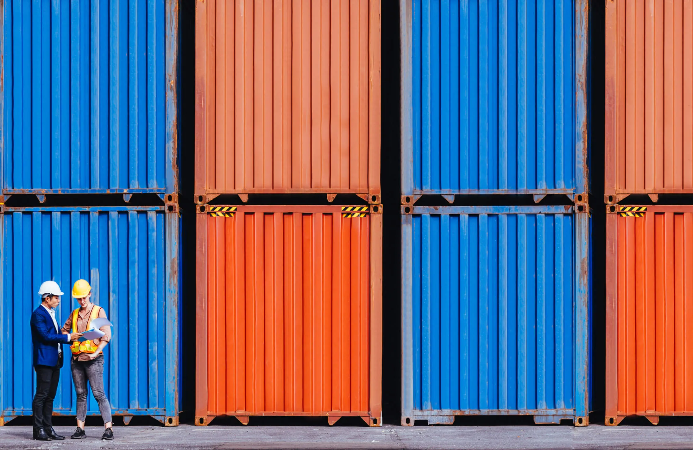Two workers in hard hats review documents in front of stacked shipping containers.