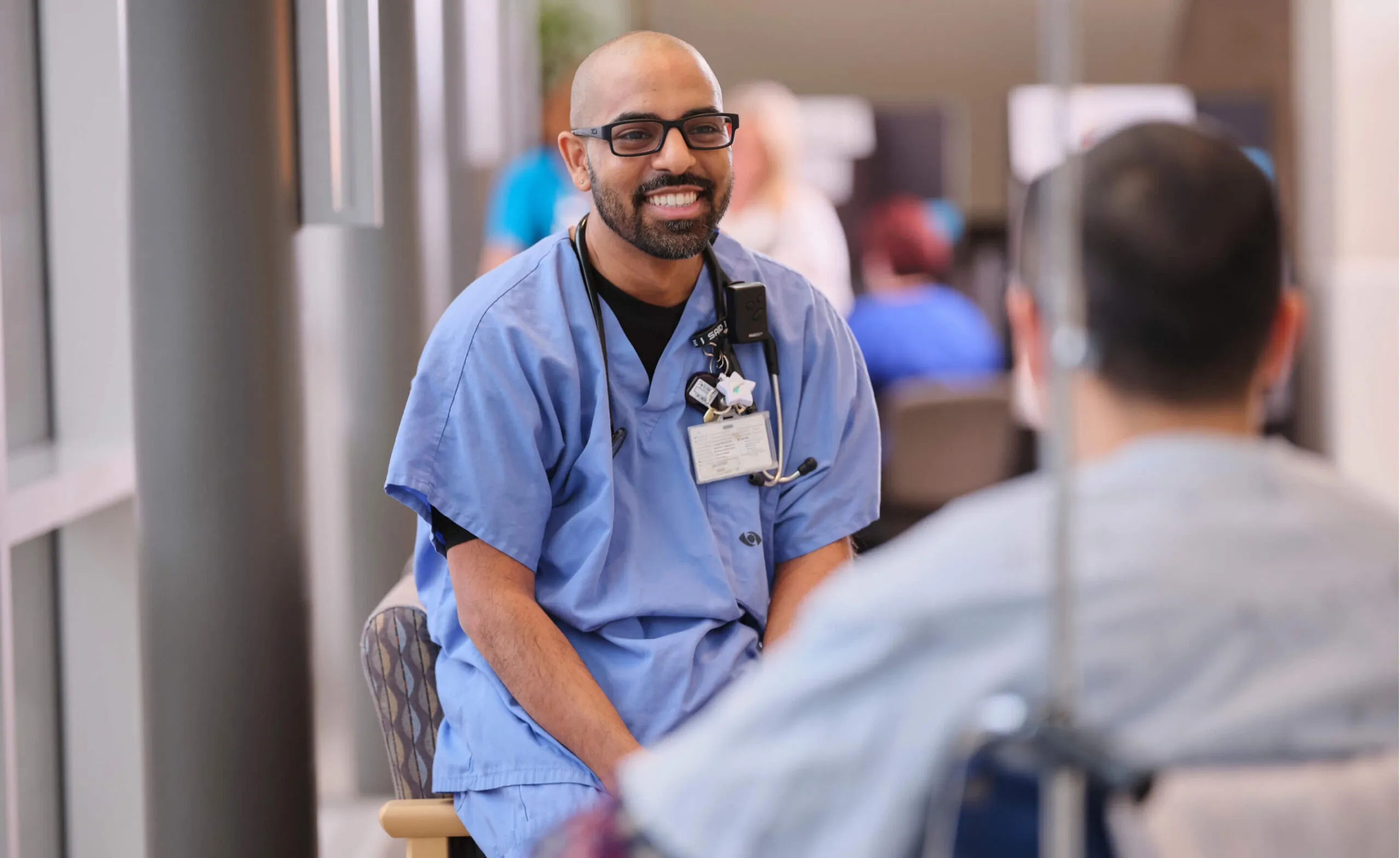 A smiling doctor in blue scrubs talks to a patient in a hospital setting.