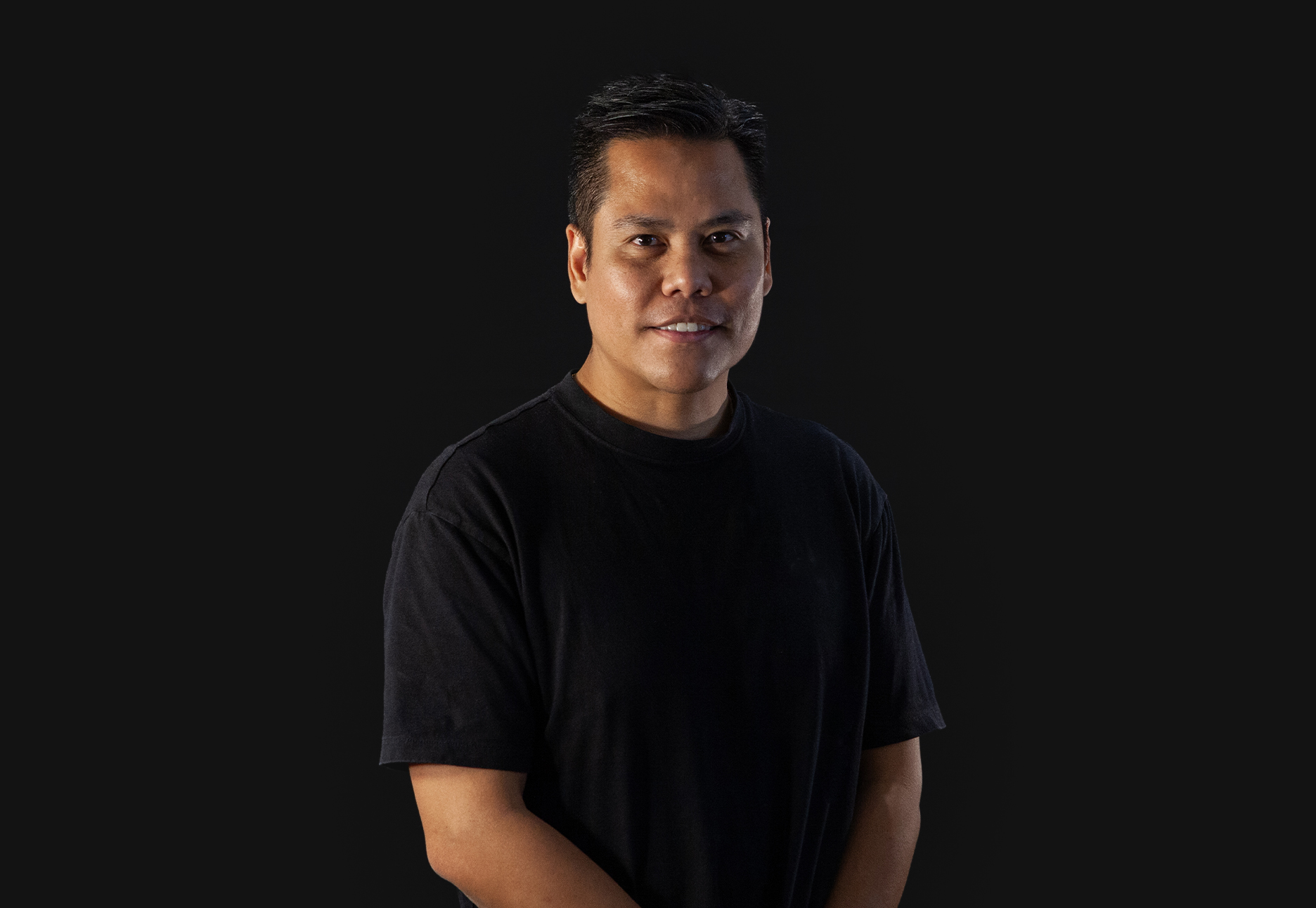 A man in a black t-shirt smiles at the camera against a black background.