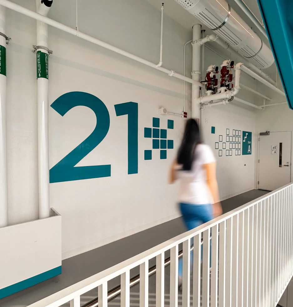 A person walks past a large number 21 on a white wall with pipes overhead.