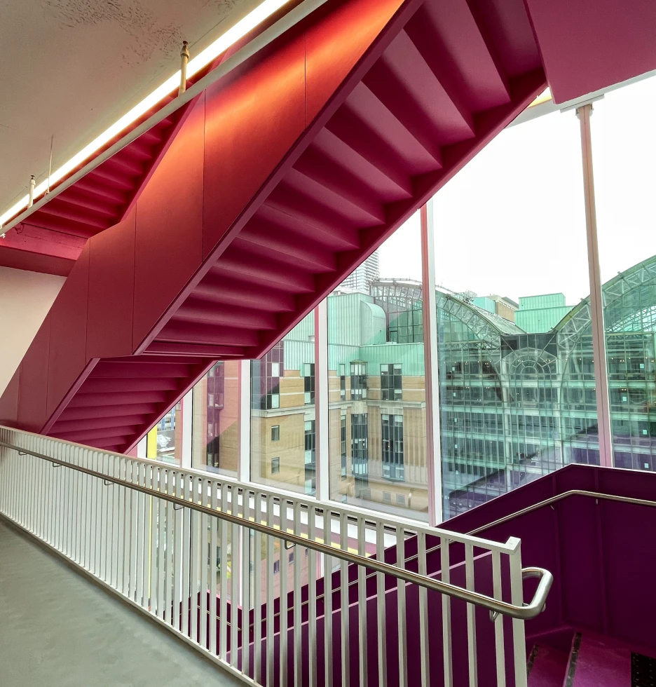 Modern pink staircase with a view of city buildings through a large window.